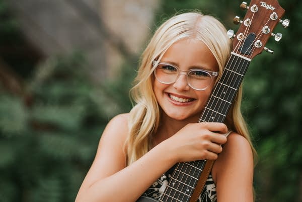 A musician sits with a guitar on a stairway near a building covered in vines.
