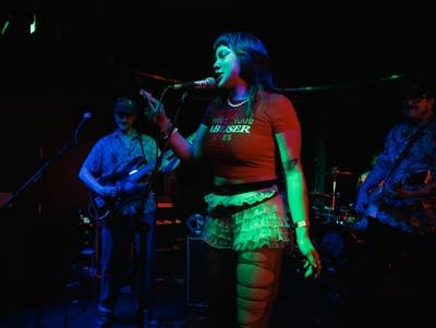 Sophie Hiroko sings into a microphone with a band playing behind her.