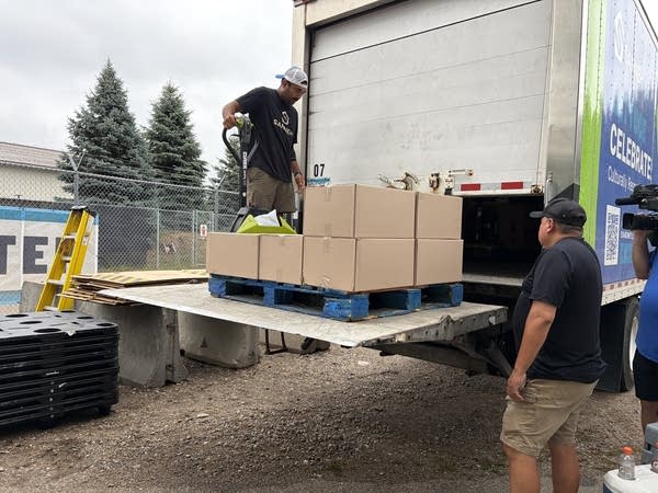 Two workers raise a palette of cardboard boxes on a cargo truck lift.