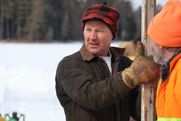 A man in a brown coat and red checkered hat holds a large plank of wood.