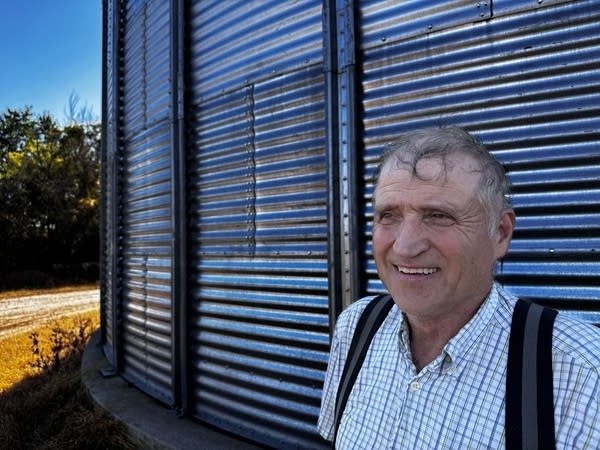 A man in a white shirt smiles in front of metal-sided grain bins.