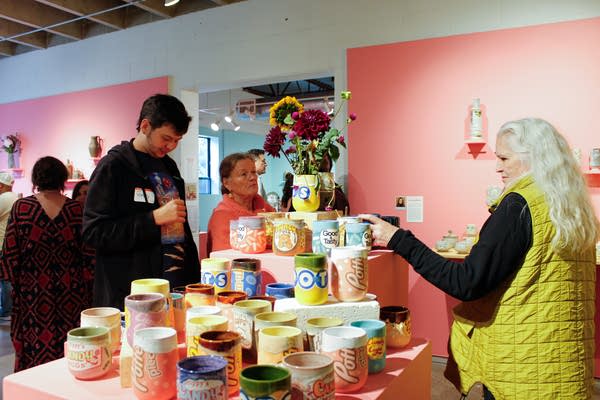 A group of people look over items of pottery on a platform in a room with pink walls and tables.