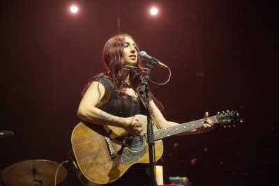 A woman sings and plays guitar onstage in a large music venue
