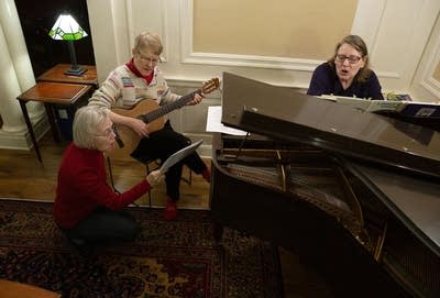 A woman plays guitar as two other people sing around a piano.