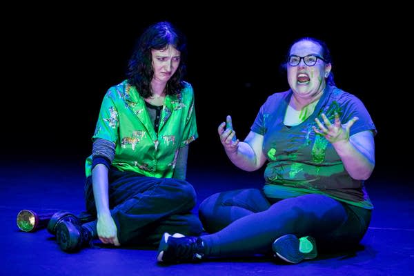 Two actors sit on a stage, one holds hands forward and yells, with slime-like substance on her clothes.