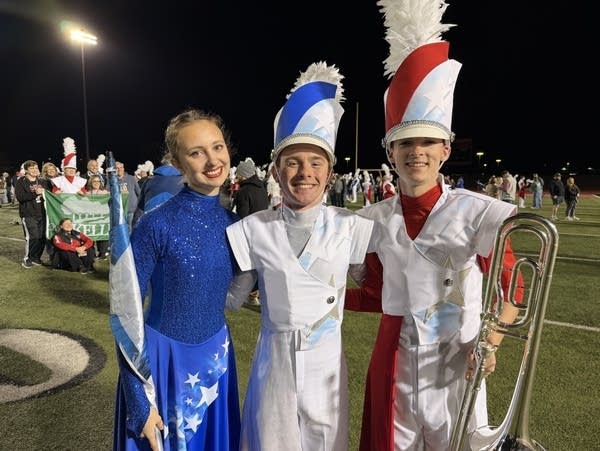 Three high school marching band players pose for a photo while wearing blue, white and red uniforms.