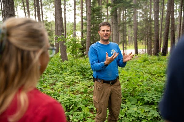 A guide in a blue shirt talks to reporters in the middle of a forest.
